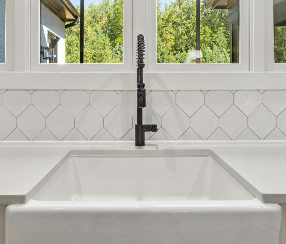 White farmhouse sink with black faucet, white backsplash with diamond-shaped tiles, and window with trees outside.