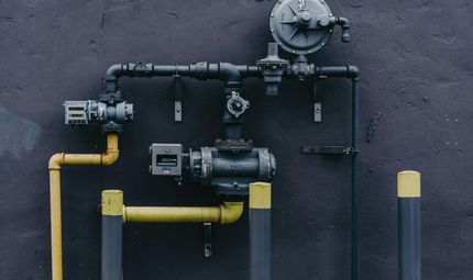 A close-up of dark-colored gas pipes, valves, and a regulator mounted on a dark wall, with some sections painted yellow.