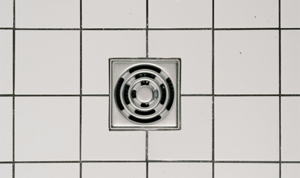 A square, chrome metal floor drain centered among white, square tiles with dark grey grout lines.