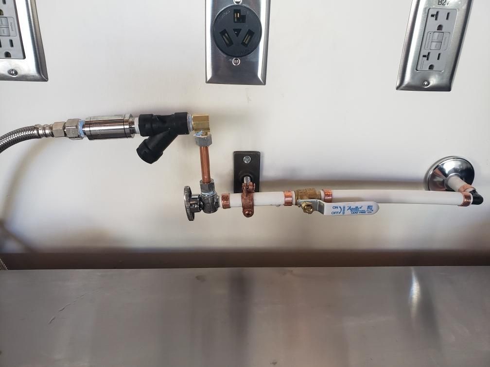 Plumbing pipes with valves and water supply line near an electrical outlet and stainless steel sink.