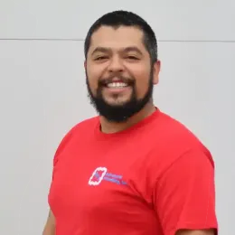 A smiling person with a beard wearing a red t-shirt with a company logo, standing against a plain white wall.