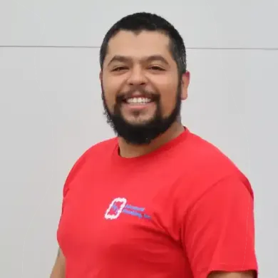 A smiling person with a beard wearing a red t-shirt with a company logo, standing against a plain white wall.