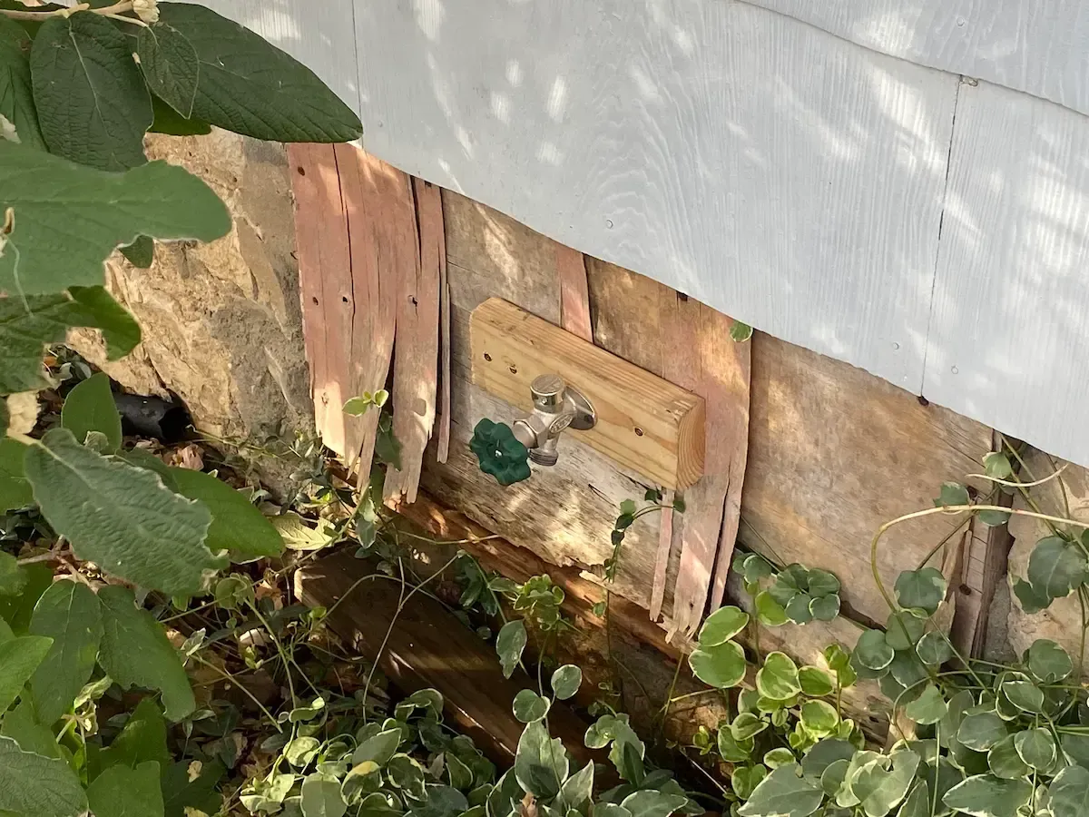 Stone foundation of a house with attached wooden structure, surrounded by plants.