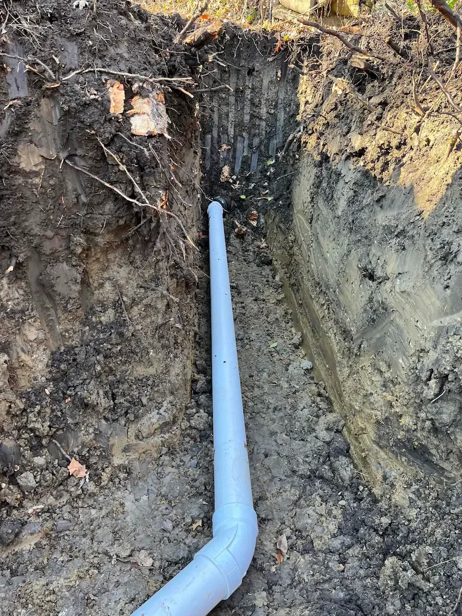 A white perforated drain pipe in a dirt trench, possibly for drainage, with soil and tree roots visible.