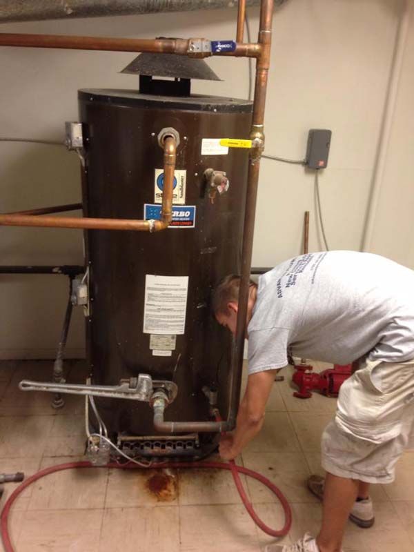 Plumber working on a brown water heater in a utility room; copper pipes, red hose.