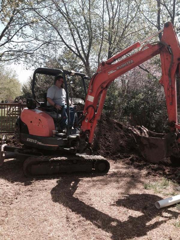 Man operating an orange excavator, digging in a yard; sunny day.