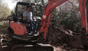 A person operates an orange compact excavator, digging into a mound of dirt in a yard.