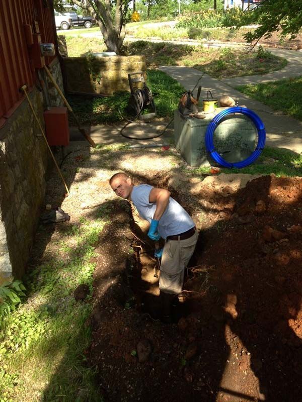 Person digging a trench next to a building, blue gloves, sunny day.