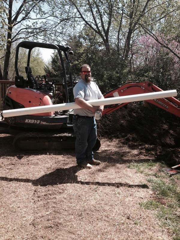 Man holding a long white pipe stands near an orange excavator and a freshly dug hole.