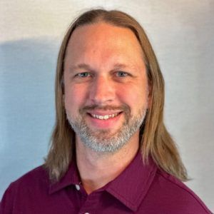 A man with long hair and a beard is wearing a purple shirt and smiling.