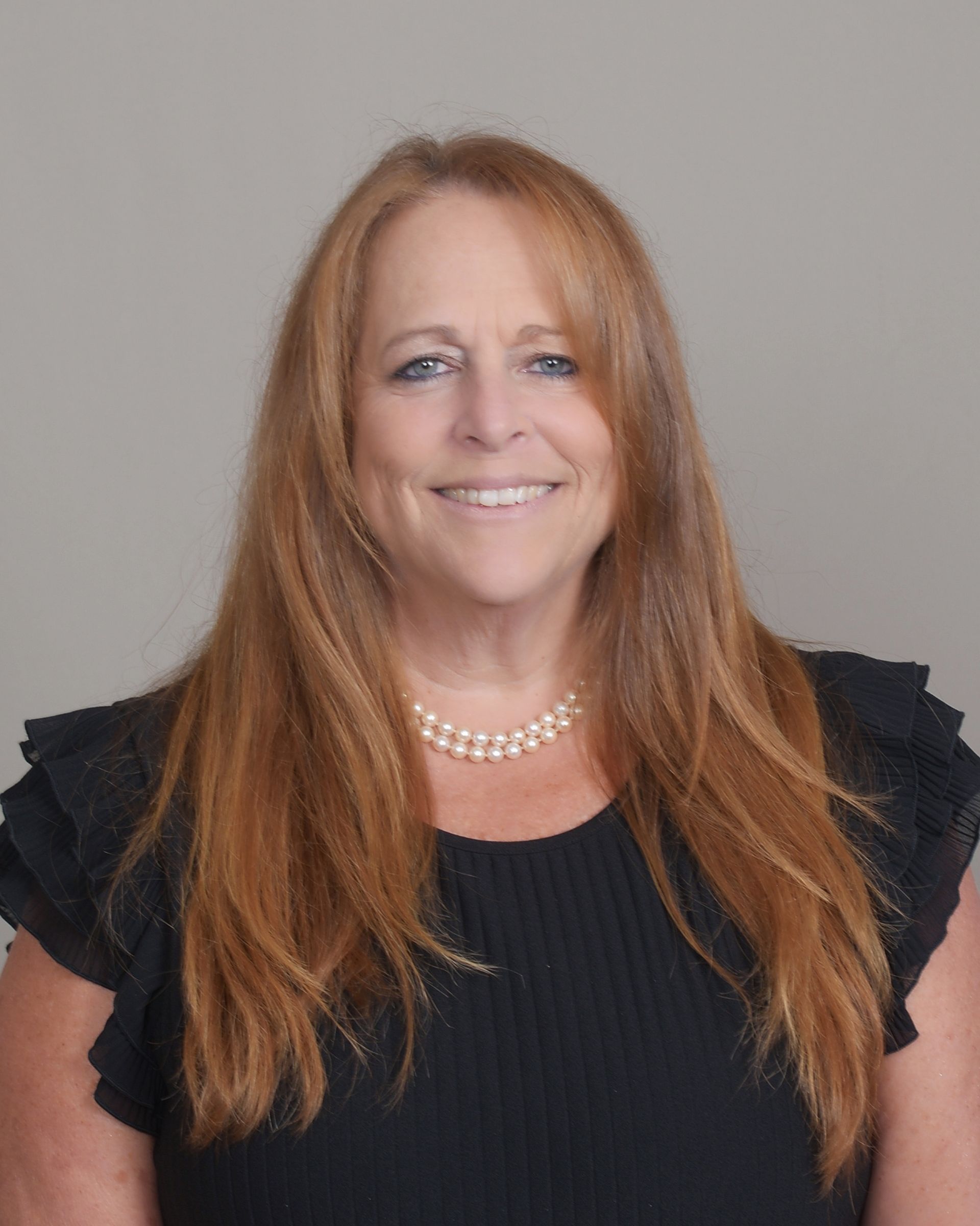 A woman with long red hair is wearing a black shirt and a pearl necklace.