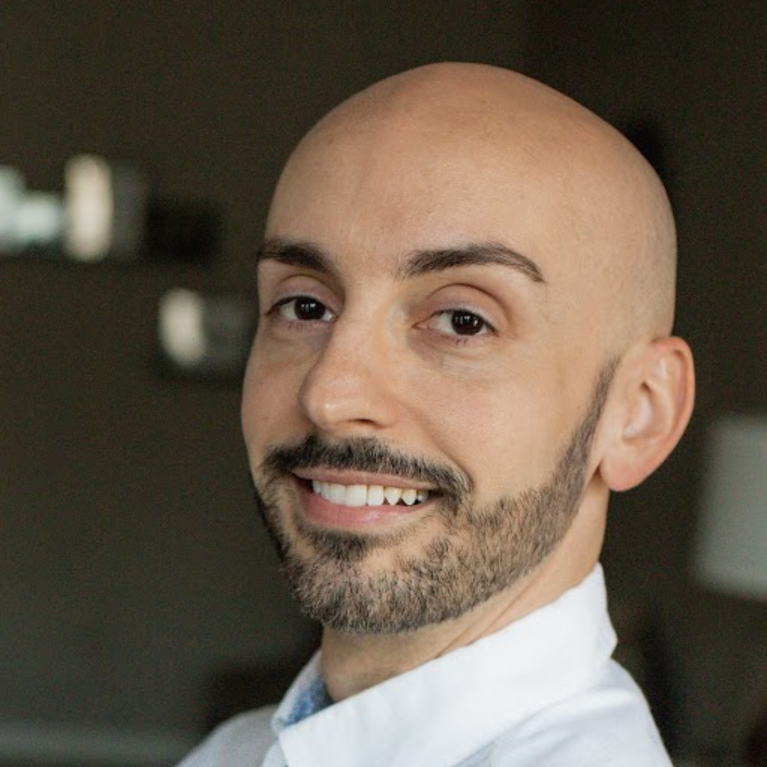 A bald man with a beard is wearing a blue suit and red tie.