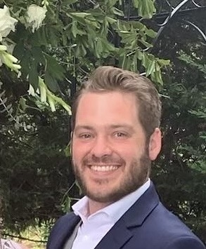 A man with a beard is smiling for the camera while wearing a blue hoodie.