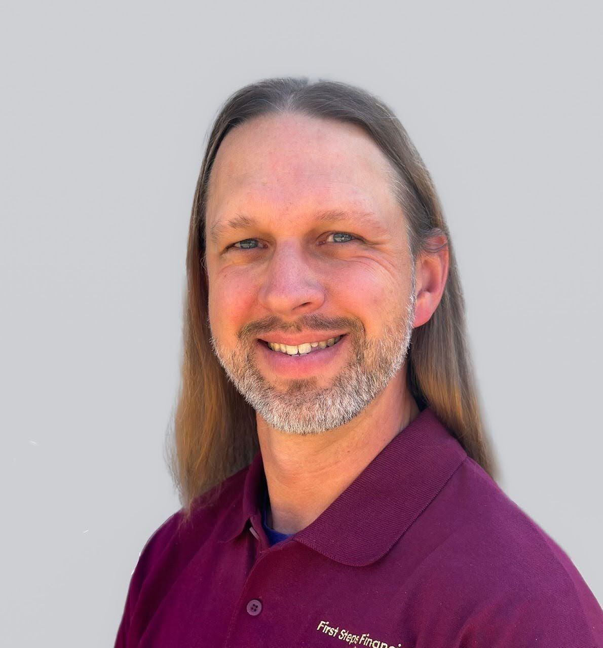 A man with long hair and a beard is wearing a purple shirt and smiling.
