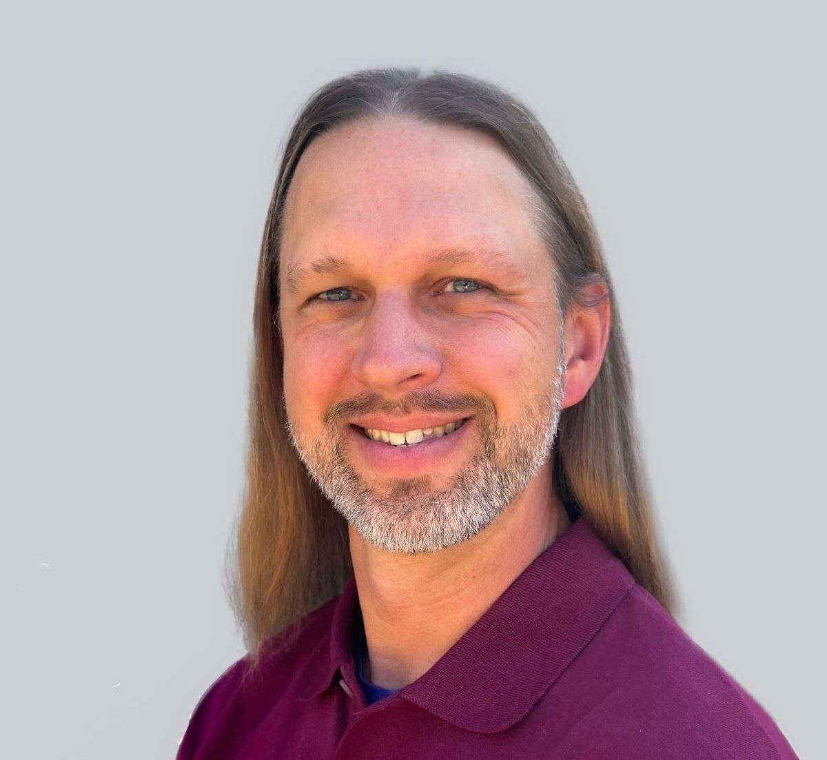 A man with long hair and a beard is wearing a purple shirt and smiling.