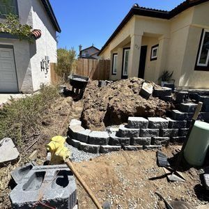 A house under construction. A retaining wall of gray stone is being built, dirt pile and tools present.