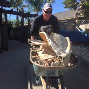 Man pushing a wheelbarrow filled with rubble on a sunny patio.