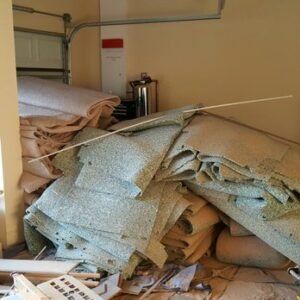 Pile of discarded carpet and padding in a garage. Beige and green colors. Garage door and fire extinguisher are visible.