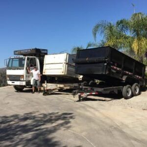 A white truck pulling two dump trailers on a sunny road. A person stands beside the truck.
