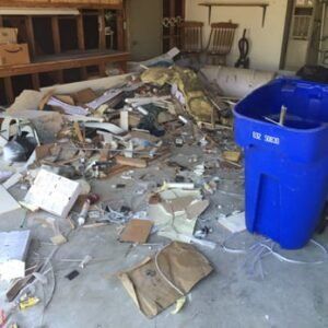 A pile of debris, including wood and insulation, on a concrete floor near a blue trash bin.