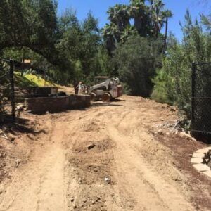 Dirt road with a small tractor and two people clearing land. Trees surround the area.