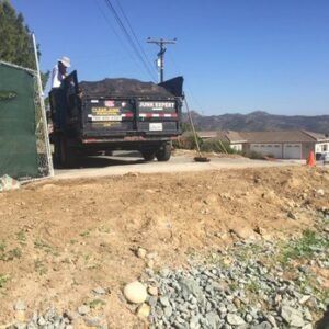 A junk removal truck parked by a gate, loading debris on a dirt road near houses and hills.