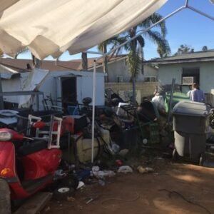 Backyard cluttered with debris, a red scooter, trash cans, and a tarp. A person stands near a building.