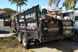 Black junk removal trailer filled with debris, parked on a curb. Phone number visible on the side.