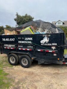 Black junk removal trailer filled with debris, parked outdoors. Phone number on side.
