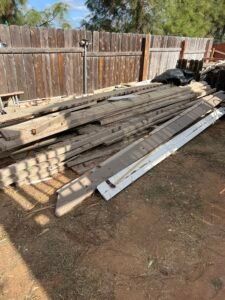 Pile of weathered wooden planks and trim on dry, grassy ground, with a wooden fence in the background.