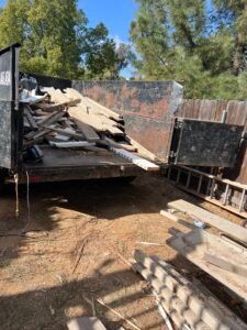 A dumpster overflowing with wood debris, likely construction waste, sits outdoors. A ladder is nearby.