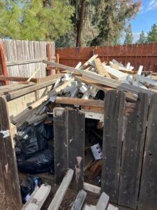 Pile of wood and trash inside a wooden fenced enclosure. Black trash bags are visible.