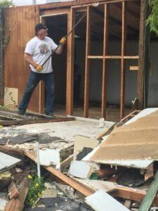 Man demolishing a wooden structure with a tool, rubble in the foreground, sunny day.