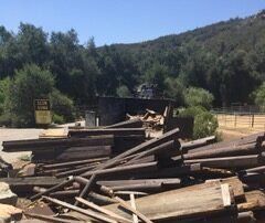 Piles of wood in a parking area with a building and trees in the background. A sign is visible.
