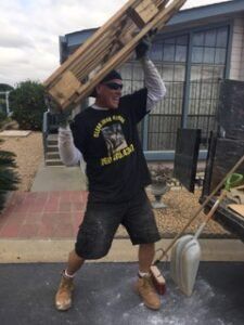 Man lifting a wooden pallet above his head outside. He wears a black shirt, shorts, and work boots.