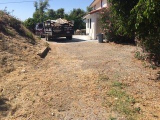 Gravel driveway leading to a light-colored house. A truck loaded with debris is parked in the driveway.