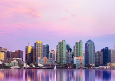 Skyline of San Diego reflecting in water at sunset; pink and orange hues.