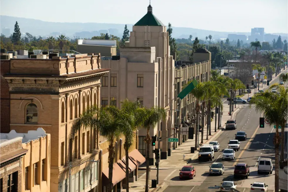 City street lined with buildings and palm trees; cars travel on the road under a bright sky.