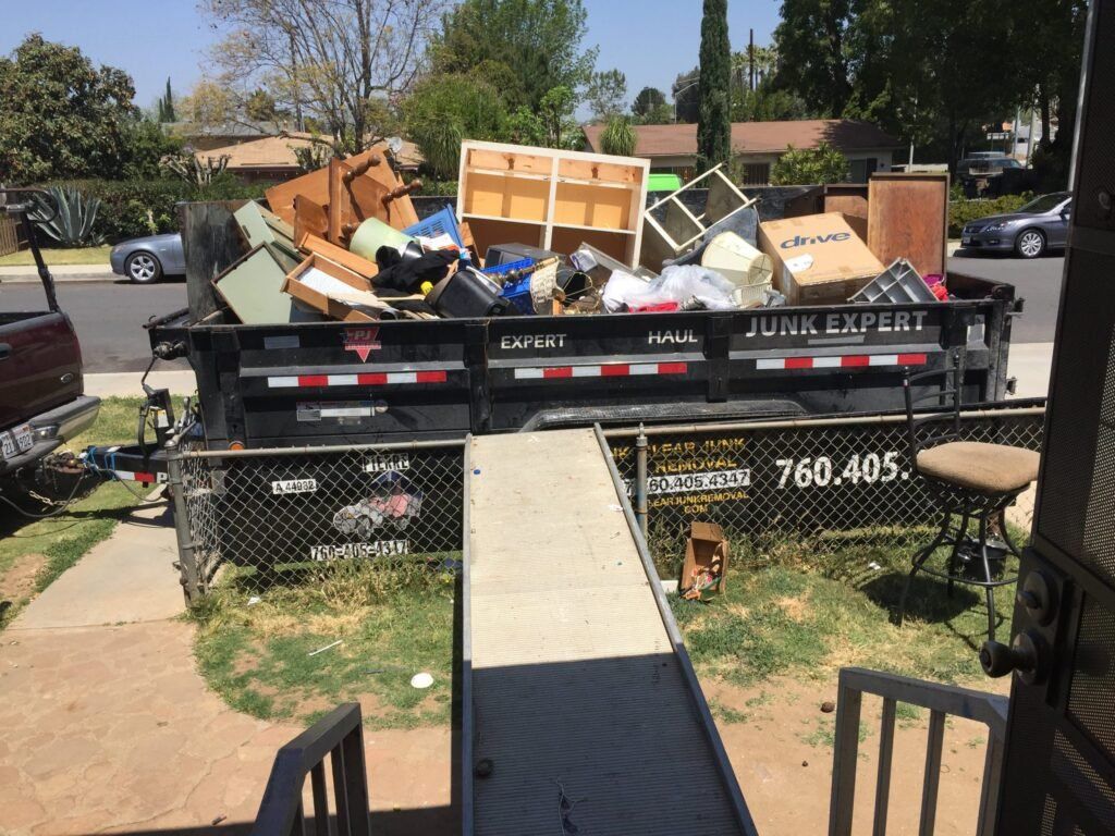 A junk-filled dumpster on a driveway, filled with furniture and debris. A ramp leads to the dumpster.