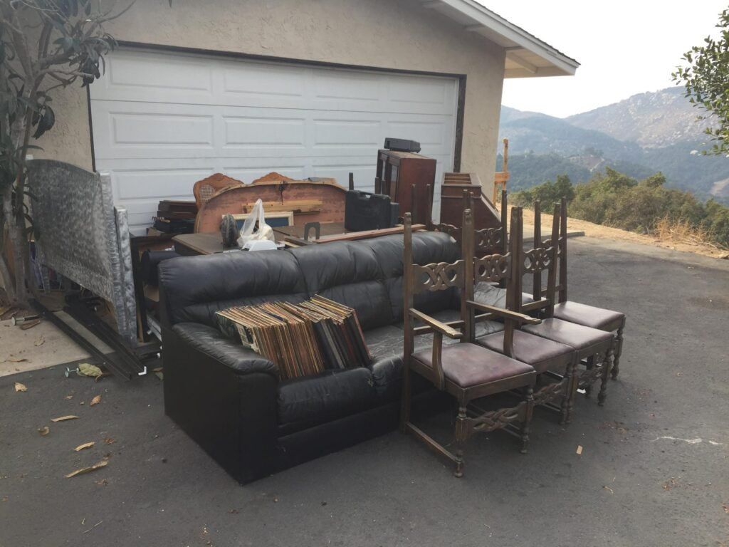 Garage door with furniture piled outside; includes sofa, chairs, and other items. Hillside in background.