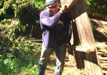 Man in blue shirt, carrying a wooden fence section outdoors. Sunny setting, green foliage.