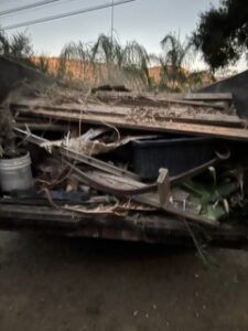 Truck bed filled with debris, including wood planks, a bucket, and yard waste, outdoors.