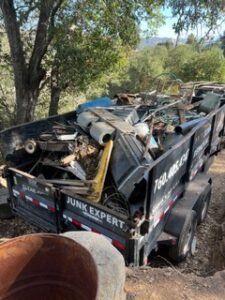 A dumpster trailer overflowing with junk, parked outdoors near trees.