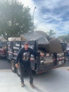 Man standing in front of a trailer filled with junk, under a cloudy sky. The trailer has 