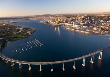 Bridge spanning blue water, connecting waterfront city and suburban area, with sailboats and buildings.