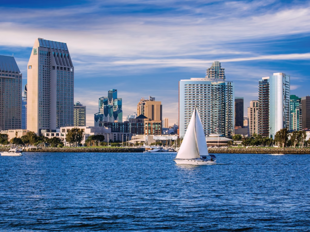 Sailboat on water in front of a San Diego skyline, blue sky and water.