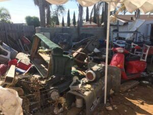 Pile of junk and debris in a yard; includes a red scooter, metal scraps, and an overturned green seat.