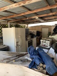 Cluttered outdoor storage area with a refrigerator, appliance, and other items under a corrugated metal roof.