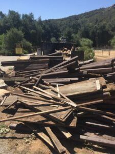 Pile of weathered wooden planks outdoors, with trees and building in the background.