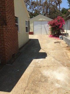 Concrete driveway next to a brick building and a cream-colored house, leading to a white garage with pink flowers.